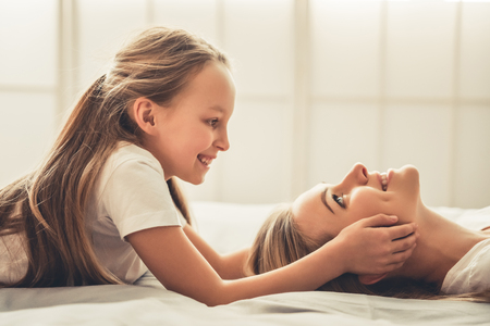Beautiful woman and her cute little daughter are smiling while spending time together on bedの写真素材