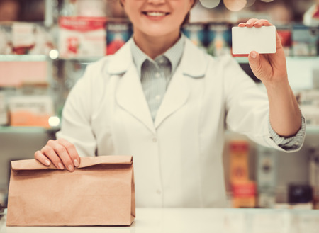 Beautiful pharmacist is smiling, holding a purchase and a card while working at the cash desk in pharmacyの写真素材