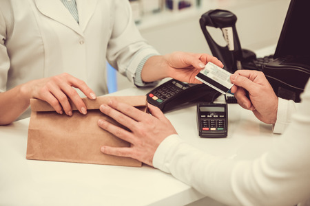 Cropped image of pharmacist giving a purchase to a client and talking a credit card while working at...