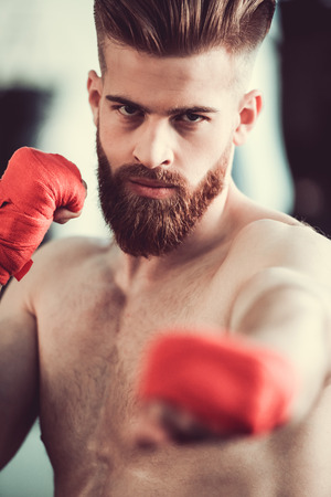 Handsome bearded boxer with bare torso is looking at camera while practicing punches at the fight clubの写真素材