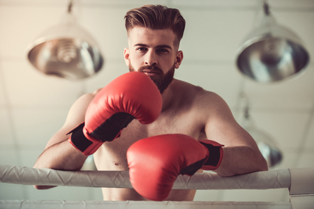 Handsome bearded boxer with bare torso in red gloves is looking at camera while standing on the boxing ringの写真素材