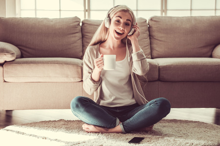 Attractive young girl in headphones is listening to music, drinking tea and smiling while sitting on the floor at homeの写真素材
