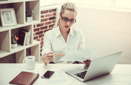 Attractive young business lady in eyeglasses is studying documents and using a laptop while working in officeの写真素材