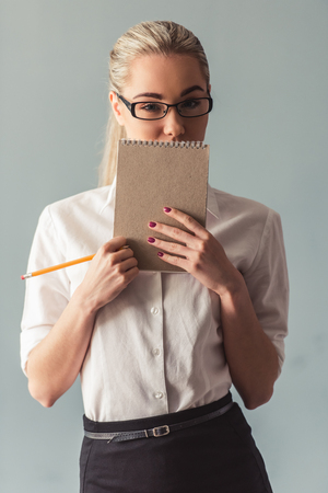 Attractive young business lady in suit is holding a notepad and a pencil and looking at camera, on gray backgroundの写真素材