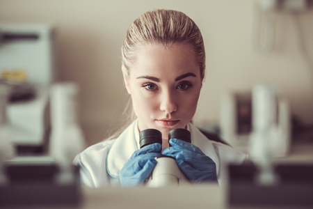 Beautiful female medical doctor is using a microscope and looking at camera while working at the labの写真素材