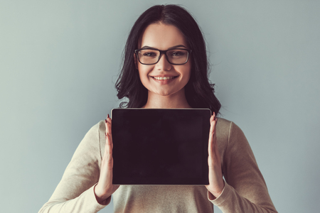 Beautiful young woman is showing a digital tablet, looking at camera and smiling, on gray backgroundの写真素材