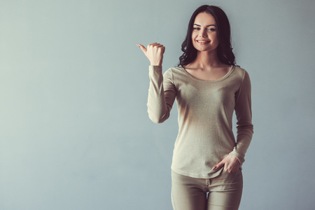 Beautiful young woman is pointing to the side, looking at camera and smiling, on gray backgroundの写真素材