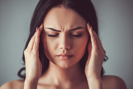 Portrait of beautiful young brunette with bare shoulders touching her temples feeling stress, on gray backgroundの写真素材