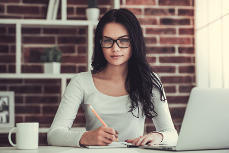 Beautiful young businesswoman in casual clothes and eyeglasses is making notes and looking at camera while working in officeの写真素材