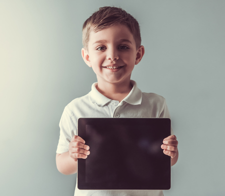 Cute little boy is holding a digital tablet, looking at camera and smiling, on gray backgroundの写真素材
