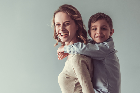 Beautiful woman and her cute little son are looking at camera and smiling, on gray background. Boy is pickabackの写真素材
