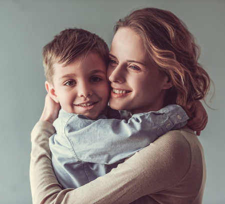 Beautiful woman and her cute little son are hugging and smiling, on gray background. Boy is looking at cameraの写真素材