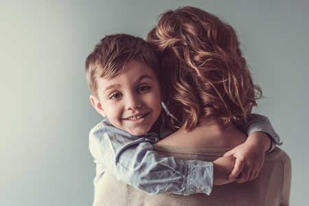 Beautiful woman and her cute little son are hugging and smiling, on gray background. Boy is looking at cameraの写真素材