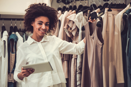 Beautiful Afro American sales assistant is using a digital tablet, looking at camera and smiling while working in female boutiqueの写真素材
