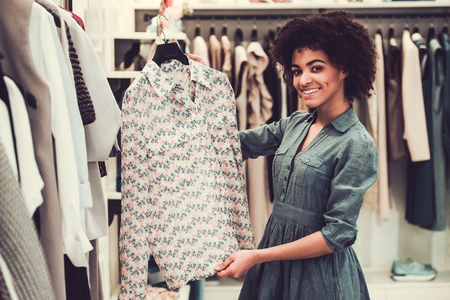 Beautiful Afro American girl is looking at camera, choosing clothes and smiling while doing shopping in female boutiqueの写真素材