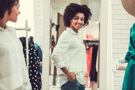 Beautiful Afro American girl is fitting on blouse, looking into the mirror and smiling while doing shopping in female boutiqueの写真素材
