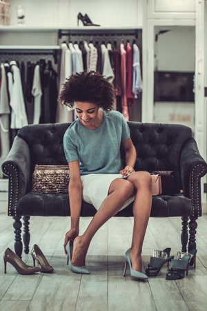 Beautiful Afro American girl is choosing high-heeled shoes and smiling while doing shopping in female boutiqueの写真素材