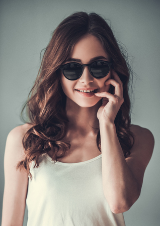 Beautiful young girl in sun glasses is looking at camera and smiling, on gray backgroundの写真素材