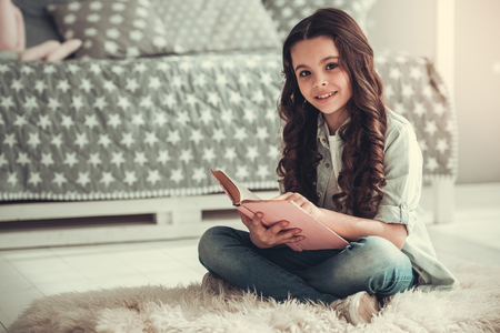 Beautiful school girl is reading a book, looking at camera and smiling while sitting in her roomの写真素材