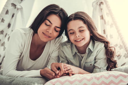 Beautiful school girl and her mom are smiling while lying in kids' tent at homeの写真素材