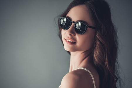 Beautiful young girl in sun glasses is looking at camera and smiling, on gray backgroundの写真素材