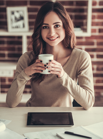 Beautiful young female student is holding a cup, looking at camera and smiling while sitting at desk at homeの写真素材