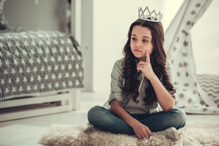 Beautiful school girl is looking away and thinking while sitting with a crown on her head in her roomの写真素材