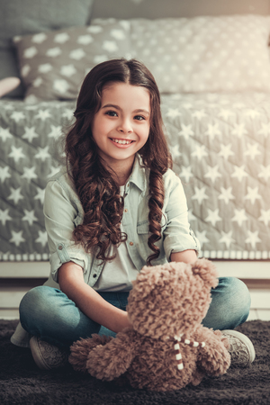 Beautiful school girl is holding a teddy bear, looking at camera and smiling while sitting in her roomの写真素材