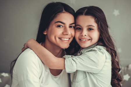 Beautiful school girl and her mom are hugging, looking at camera and smiling while sitting in girl's room at homeの写真素材