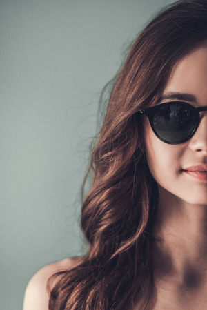 Cropped image of beautiful young girl in sun glasses looking at camera and smiling, on gray backgroundの写真素材