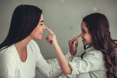 Beautiful school girl and her mom are touching their noses and smiling while playing in girl's room at homeの写真素材