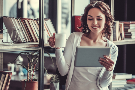 Portrait of a young beautiful girl using tablet at the shelves of books in the home.の写真素材