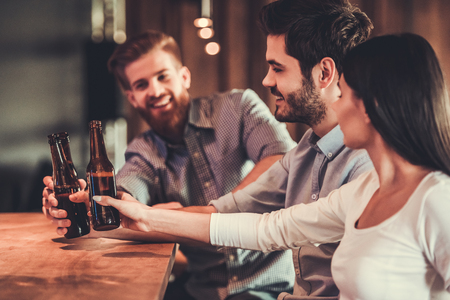 Happy young friends in casual clothes are  smiling and clinking bottles of beer while sitting at bar counter in pub.の写真素材