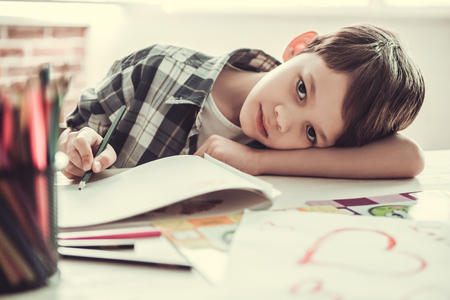 Handsome schoolboy is looking at camera and leaning on the table while drawing in his room at homeの写真素材