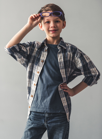 Stylish little boy in casual clothes and glasses is looking at camera and smiling, on gray backgroundの写真素材