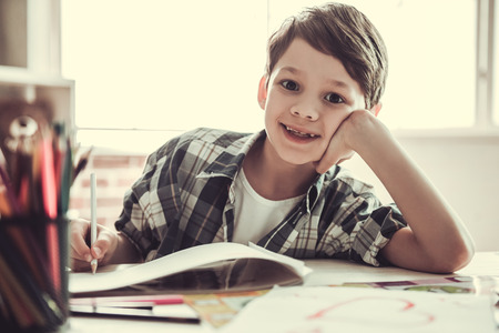 Handsome schoolboy is looking at camera and smiling while drawing in his room at homeの写真素材