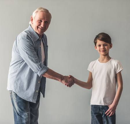 Handsome grandpa and grandson are shaking hands, looking at camera and smiling, on gray backgroundの写真素材