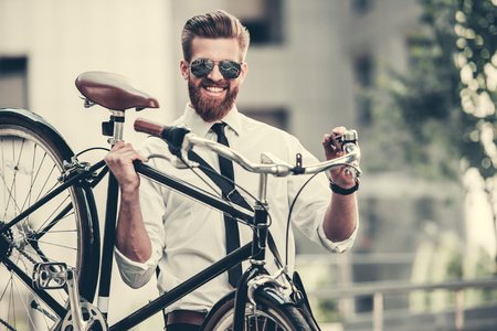 Handsome bearded businessman in classic suit and sun glasses is carrying his bicycle and smilingの写真素材