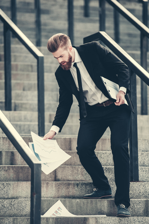 Handsome bearded businessman in classic suit is dropping documents while going down the stairs in the city centerの写真素材