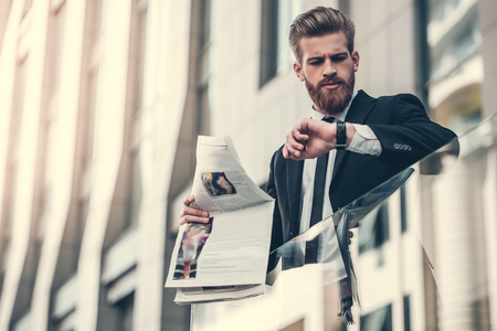 Handsome bearded businessman in classic suit is holding a newspaper and looking at his watch while standing on the balcony of the office centerの写真素材