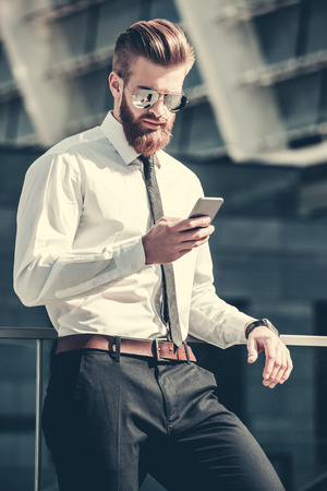 Handsome bearded businessman in classic suit and sun glasses is using a smart phone while standing on the balcony of the office centerの写真素材