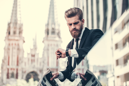 Handsome bearded businessman in classic suit is holding a newspaper and looking at his watch while standing on the balcony of the office centerの写真素材