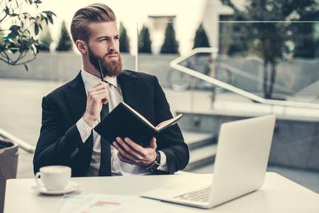 Handsome bearded businessman in classic suit is using a laptop and taking notes while sitting in cafe in the city centerの写真素材