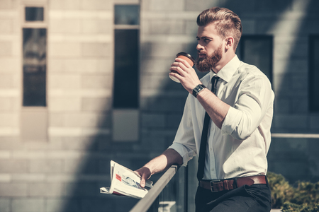 Handsome bearded businessman in classic suit is drinking coffee and holding a newspaper while resting on the balcony of the office centerの写真素材