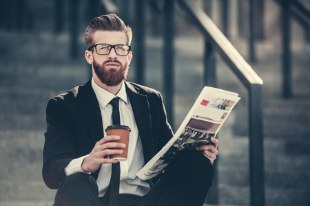 Handsome bearded businessman in classic suit and eyeglasses is holding a cup of coffee and reading newspaper while sitting on the stairs in the city centerの写真素材