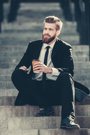 Handsome bearded businessman in classic suit is holding a cup of coffee and looking away while sitting on the stairs in the city centerの写真素材