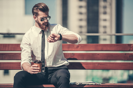 Handsome bearded businessman in classic suit and sun glasses is drinking coffee and looking at his watch while resting on the bench in the city centerの写真素材