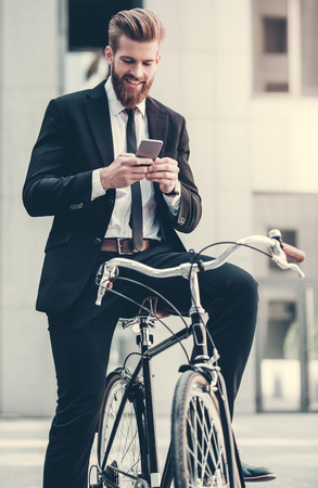 Handsome bearded businessman in classic suit is using a smart phone and smiling while riding bicycle in cityの写真素材