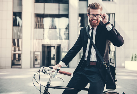 Handsome bearded businessman in classic suit and with bag is looking at camera and smiling while riding bicycle in cityの写真素材