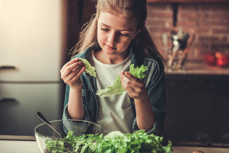 Cute teenage girl is adding lettuce into the bowl while preparing salad in kitchenの写真素材
